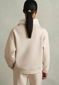 Světle béžová oversized bunda s vysokým límcem a žebrovanými manžetami, která má viditelný šev a hladkou texturu látky.