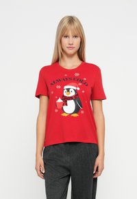 Blond kvinde iført rød T-shirt med pingvin i nissehue og tørklæde, der holder en varm drik, ledsaget af teksten "Alltid kold" og snefnug.