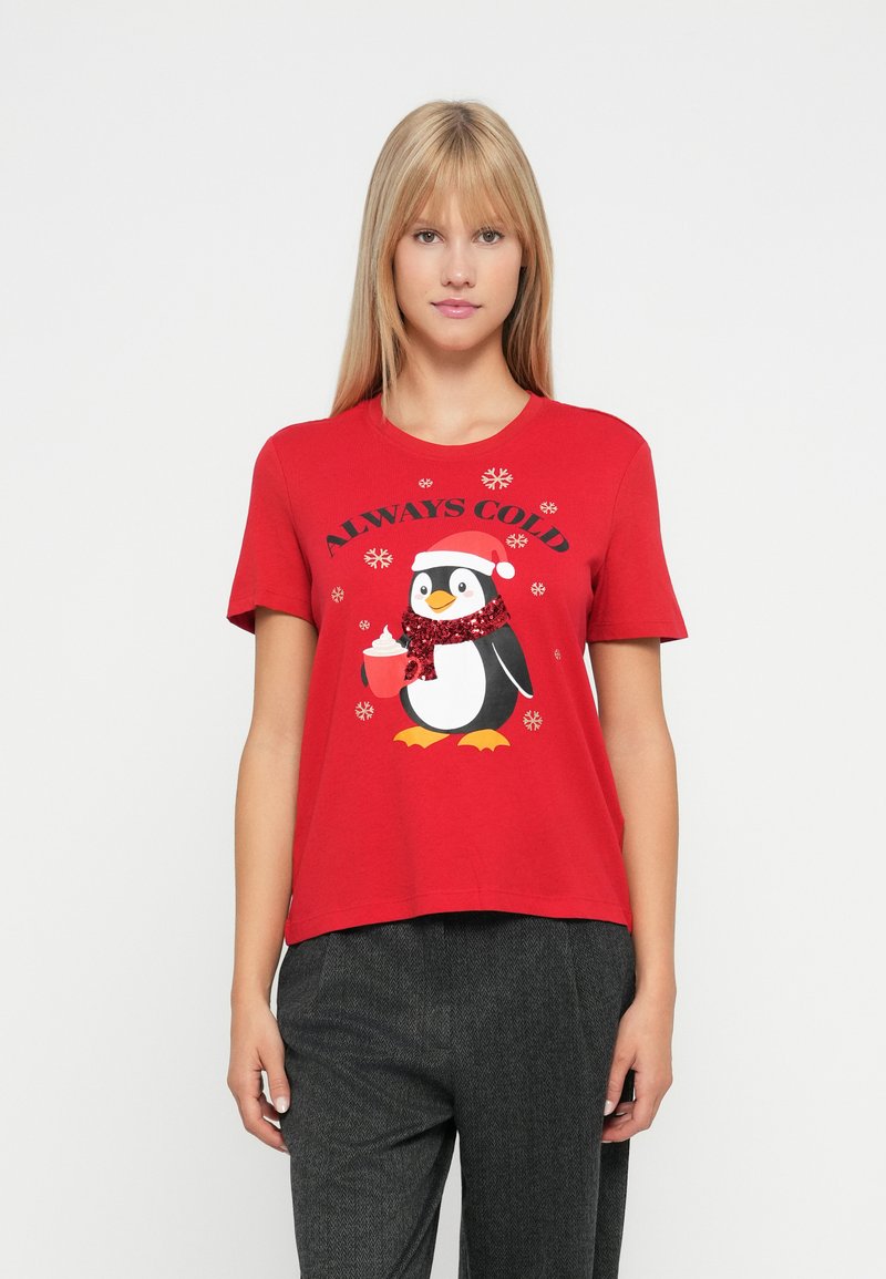 Blond kvinde iført rød T-shirt med pingvin i nissehue og tørklæde, der holder en varm drik, ledsaget af teksten "Alltid kold" og snefnug.