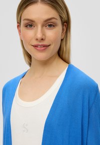 Niebieski sweter kardigan o luźnym kroju, noszony na kremowym podkoszulku z okrągłym dekoltem, ozdobionym wyhaftowanym logo. Gładka tekstura, casualowy styl.