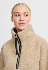 Beige fleece pullover med høj krave, lynlås og sorte lynlåsdetaljer. Teksturen er blød med et boucle-mønster.