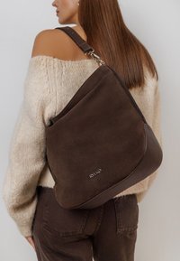 Houten schoudertas van bruine suede met een gestructureerde leren bodem, voorzien van een zilveren logo-detail en een verstelbare schouderband. Brede lichaamsontwerp.