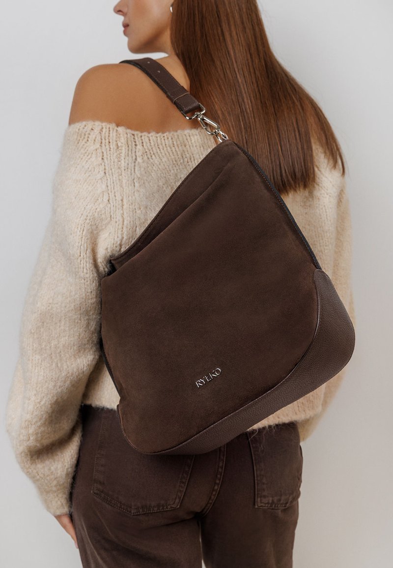 Houten schoudertas van bruine suede met een gestructureerde leren bodem, voorzien van een zilveren logo-detail en een verstelbare schouderband. Brede lichaamsontwerp.