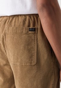 Pantalone od kordura svijetlosmeđe boje s teksturiranom površinom, s jednim stražnjim džepom i malom crnom etiketom blizu struka.