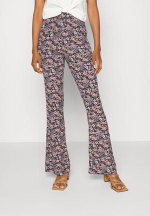Persona con pantalones negros de tiro alto con un patrón floral colorido y sandalias de tacón marrones con tiras, combinados con un top blanco anudado.