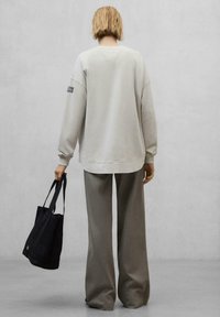 Sweatshirt oversized gris clair avec des poignets côtelés, associé à un pantalon large gris. Sac fourre-tout noir tenu d'une main. Design simple et minimal.