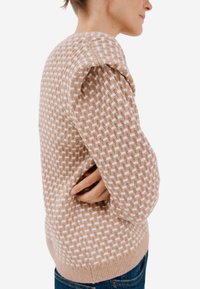 Sweter dziany w jasnobrązowym kolorze z białym wzorem w kratkę, z teksturowanym materiałem, długimi rękawami i ściągaczem na dole. Noszony z niebieskimi dżinsami.