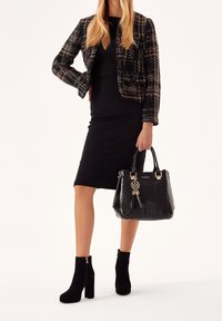 Bolso de mano texturado negro con acentos dorados y un fleco. La modelo lleva un vestido negro ajustado, una chaqueta negra de cuadros y botas negras hasta el tobillo.