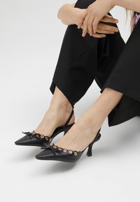 Svarta läderslingback-klackar med spetsig tå, båge och metallöglor längs kanten, med en låg stilettklack.