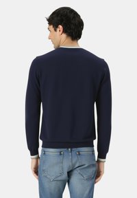 Uomo che indossa un maglione a maniche lunghe blu navy con rifiniture grigio chiaro su collo e polsini, abbinato a jeans in denim blu chiaro, di spalle.