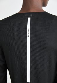 Achterkant van een persoon die een zwart mesh shirt draagt met een verticale witte strook waarop de tekst "Calvin Klein" in zwarte letters staat.