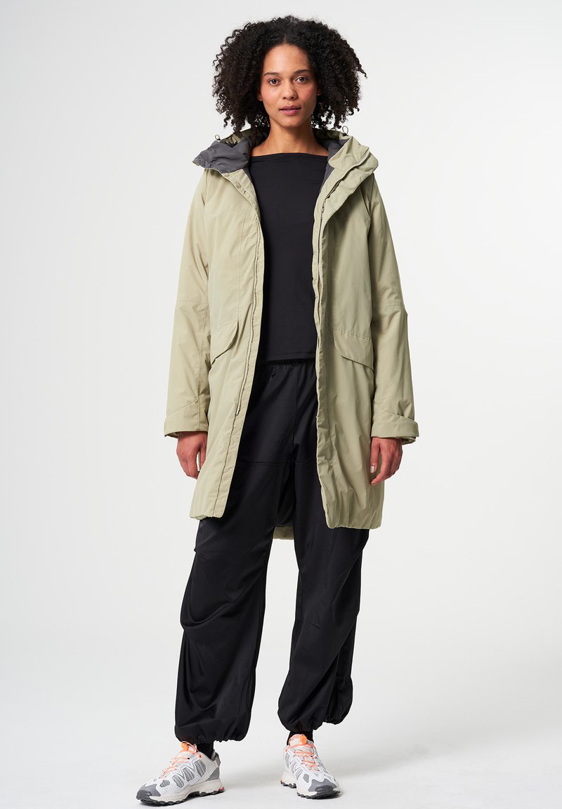 Lange, leichte khakifarbene Parka mit Kapuze, kontrastierendem grauem Innenfutter, zwei vorderen Taschen und einer lockeren Passform, kombiniert mit schwarzen Trainingshosen und Sneakers.