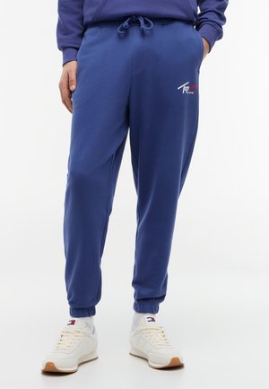 Blauwe joggerbroek met elastische boorden en een taille met trekkoord, gedragen met witte sneakers en witte sokken.