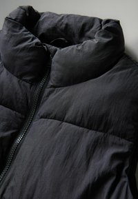 Chaleco acolchado oscuro con cuello alto, cremallera frontal y diseño acolchado con una textura ligeramente arrugada. Fabricado en tela ligera.