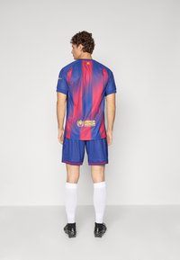 Tricou sportiv cu dungi albastre regal și roșii, cu mâneci scurte, asortat cu pantaloni scurți bleumarin. Prezintă logo-ul UNHCR pe spate.