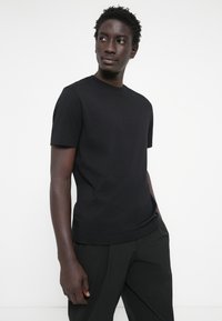 Svart bomullströja med rund halsringning och korta ärmar, slät textur och klassisk passform, bärs med mörka byxor och en neutral bakgrund.