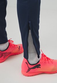 Jūras spēku sporta bikses ar rāvējslēdzēja atverēm pie pēdu daļām, kombinētas ar spilgti rozā Puma sporta apaviem ar gradientu dizainu un melnu logotipu.