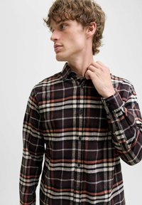 Jonge man die de kraag van een langemouwenshirt met zwart, oranje en witte ruitpatroon aanpast, terwijl hij naar links kijkt tegen een effen lichte achtergrond.