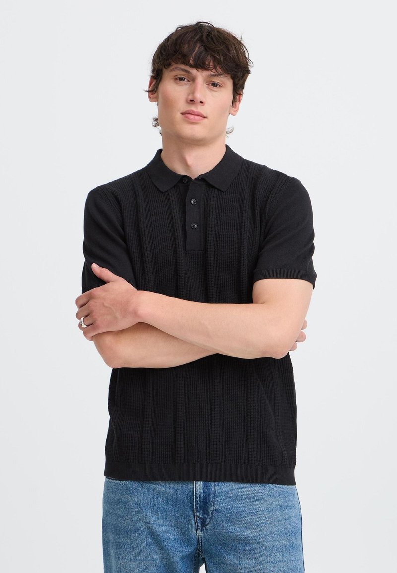 Jeune homme aux cheveux foncés portant un polo en maille noir à manches courtes et un jean bleu, debout les bras croisés devant un fond uni.