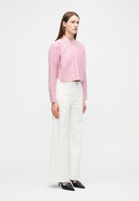 Γυναίκα που φοράει ροζ και λευκό ριγέ κοντό τοπ, λευκό παντελόνι ψηλόμεσο, με φαρδιά μπατζάκια και μαύρες μυτερές γόβες με χρυσές λεπτομέρειες.