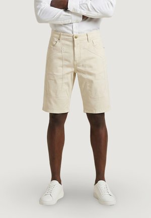 Hombre con pantalones cortos beige hasta la rodilla, camisa blanca de manga larga con los brazos cruzados y zapatillas blancas, de pie frente a un fondo liso.