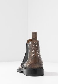 Botín de cuero marrón con laterales elásticos negros, acentos con tachuelas y un acabado texturizado. Suela plana y tira para facilitar el uso.