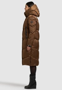 Rjava puhovka z odstranljivo kapuco, kvadratne oblike, z vezenim vzorcem in žepi z zadrgo. Oblečena s črnimi gležnjaki.