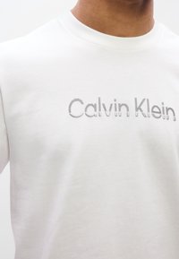 Λευκό βαμβακερό T-shirt με κεντημένο το λογότυπο "Calvin Klein" σε γκρι απόχρωση με ελαφριά ριμπ υφή, στρογγυλή λαιμόκοψη και κοντά μανίκια.