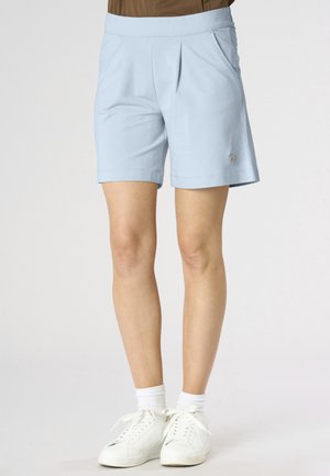 Pantaloncini azzurri chiaro fino al ginocchio con pieghe frontali, indossati con calzini bianchi e scarpe da ginnastica bianche su una persona in piedi davanti a uno sfondo semplice.