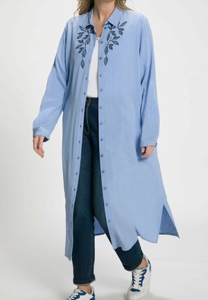 Camisa larga azul claro con botones al frente, diseño bordado de hojas en el cuello y aberturas laterales. Llevada sobre una camiseta blanca y jeans oscuros.