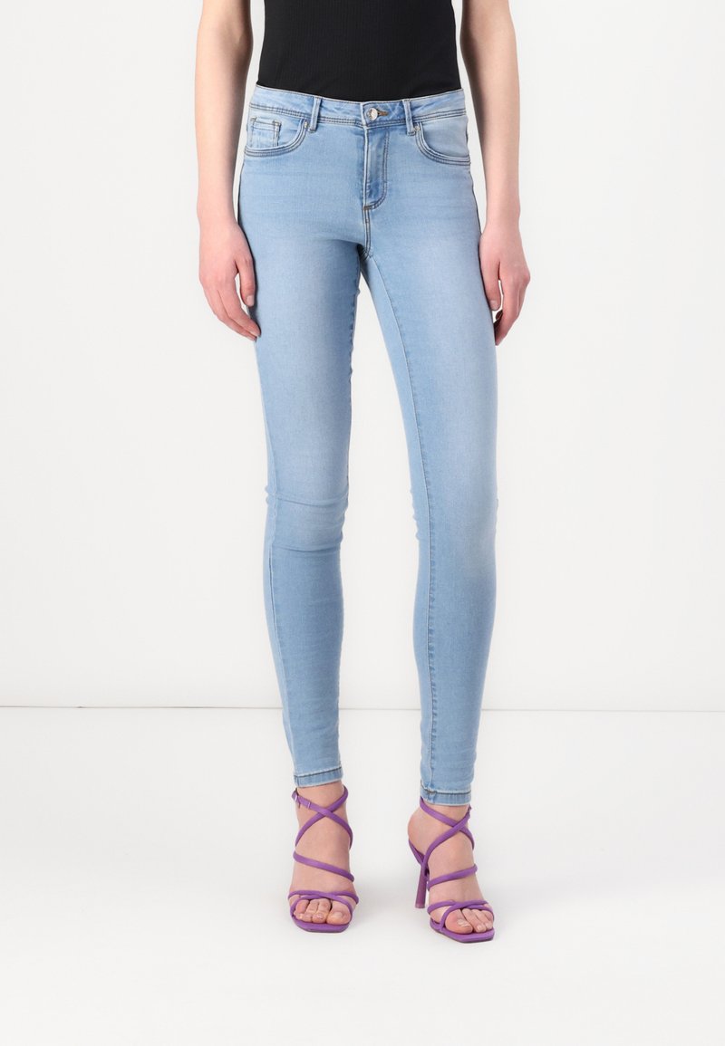 Pantalones vaqueros ajustados de color azul claro con cintura media, cuatro bolsillos y un leve desvanecimiento, combinados con sandalias de tacón alto moradas con tiras.