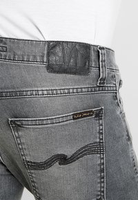 Grå denimjeans med en svart läderetikett i midjan. Har en distinkt design på bakfickan med sömdetaljer och en liten logotag.
