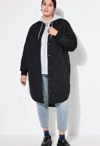 Cappotto lungo trapuntato nero con bottoni a pressione, dotato di cappuccio e vestibilità oversize, indossato sopra una felpa grigia e pantaloni azzurri chiari con motivo.