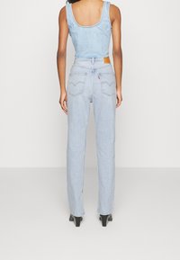 Mono vaquero de mezclilla azul claro con espalda redondeada, cuerpo ajustado y pantalón de pierna ancha. Cuenta con dos bolsillos traseros y una etiqueta de marca en tono marrón en la cintura.