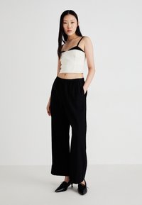 Hvit crop top med tynne svarte stropper, kombinert med svarte vide bukser. Modellen har på seg svarte hæler, som fremhever de rene linjene og det enkle designet.