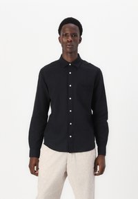 Camicia nera a maniche lunghe con bottoni, realizzata in un tessuto testurizzato, caratterizzata da una tasca sul petto e bottoni bianchi, abbinata a pantaloni chiari.
