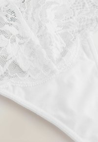 Perizoma bianco in pizzo con intricati motivi floreali e un tessuto morbido e liscio. Accentato da un delicato bordo lungo i lati.