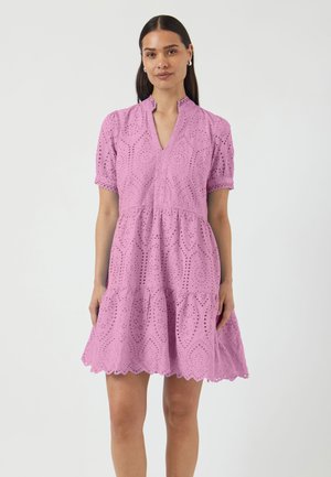 Naine kannab lühikeste varrukatega lavendlivärvi tikanditega kleiti lainelise äärega ja V-kaelusega, seisab ühetaustalise heleda tausta ees.