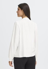 Blusa bianca a maniche lunghe con vestibilità relaxed. Presenta dettagli arricciati ai polsini e un collo arrotondato. Tessuto liscio e leggero.