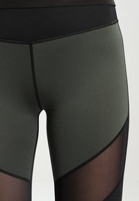 Hoge taille leggings met een donker groene hoofdfabricage, zwarte mesh-inzetstukken en contrasterende stiksels, ontworpen met een geometrisch patroon.