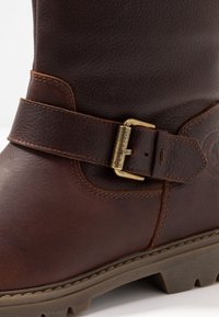 Gros plan d'une botte en cuir marron avec une bride et une boucle en laiton gravée du logo Panama Jack, avec une couture détaillée sur la semelle et la tige.
