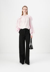 Blusa rosa chiaro con volant e maniche lunghe abbinata a pantaloni neri a vita alta. La modella tiene in mano una piccola borsa nera.