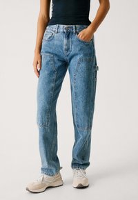 Modre ravne jeans hlače iz džinsa z več žepi, vidno šivanjem in rahlo zbledelim pranje. Ujemajo se s bež supergami.