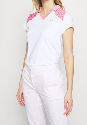 Vrouw draagt een wit polohemd met korte mouwen, met roze patroon accenten op de schouders en lichtroze broek, tegen een effen achtergrond.