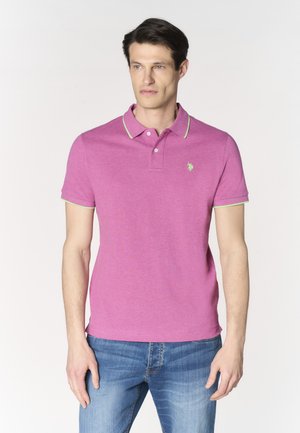 Uomo che indossa una polo viola aderente con rifiniture verdi e jeans blu, in piedi davanti a uno sfondo chiaro uniforme, guarda verso destra.