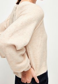 Femme portant un pull beige clair en tissu duveteux avec des manches bouffantes surdimensionnées et un jean bleu foncé, la main reposant dans la poche arrière.