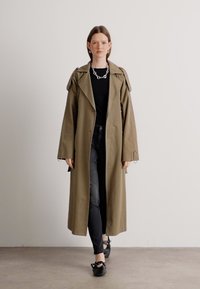 Hosszú olíva zöld trench coat, oversized hajtókákkal, fekete top és farmer párosítva. Fekete lapos cipők, masnis díszítéssel egészítik ki a megjelenést.