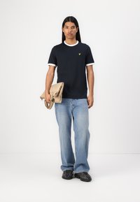 T-shirt à manches courtes bleu marine avec un col et des poignets blancs, associé à un jean bleu clair. Tenant un sac à main beige, portant des baskets noires.