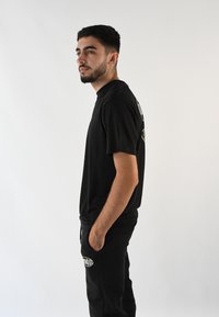 Camiseta negra de manga corta con cuello redondo. Presenta un detalle de logo en un color claro en la parte posterior. Combinada con pantalones negros que tienen un logo de acento.