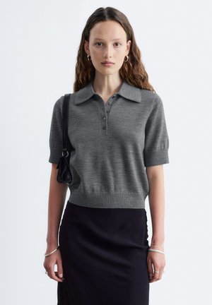 Jeune femme aux longs cheveux porte un pull gris à manches courtes, une jupe noire, des bijoux argentés et porte un sac à bandoulière noir sur fond blanc.